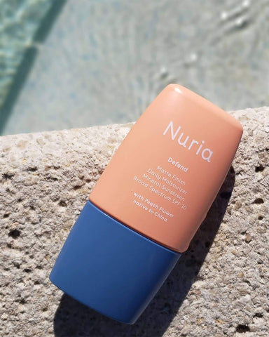 Nuria Defend Matte Finish Daily Moisturizer Mineral Sunscreen SPF 30 - bottle resting on stone with water on background